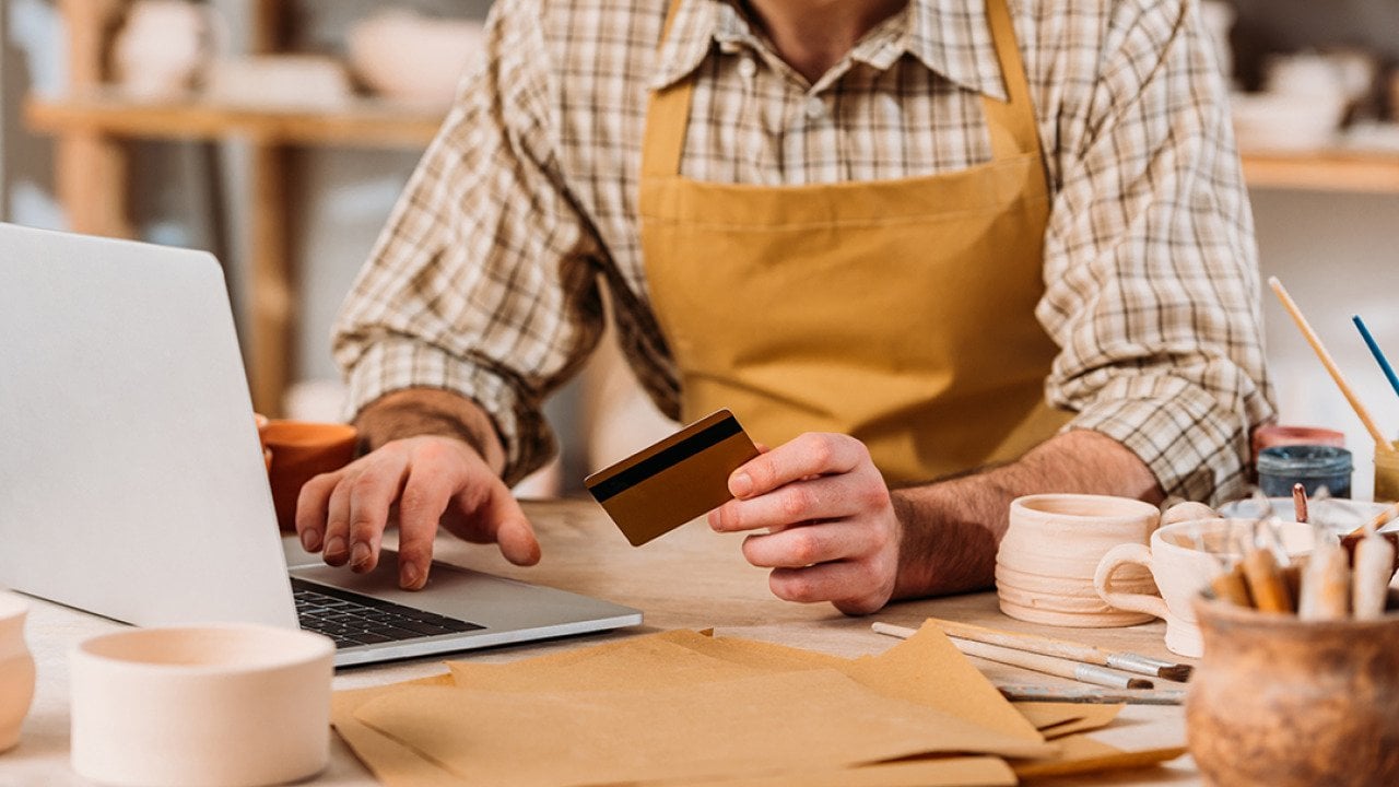 Cropped view of a small pottery business owner holding a credit card and browsing a laptop.