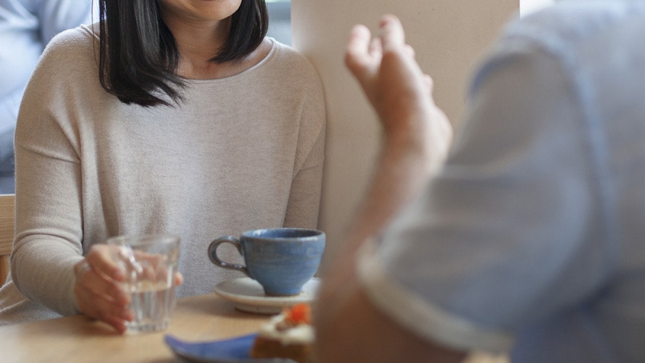 Two people sitting across from each other at a table in a cafe with water, a piece of cake, a mug in front of them. Just one person's smiling face is partially in view. 