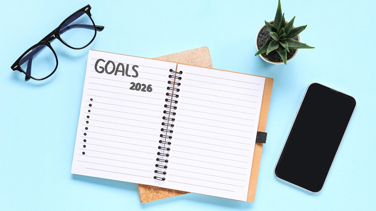 A pair of eyeglasses, a smartphone, and a notebook to list 2026 goals with a blue background.