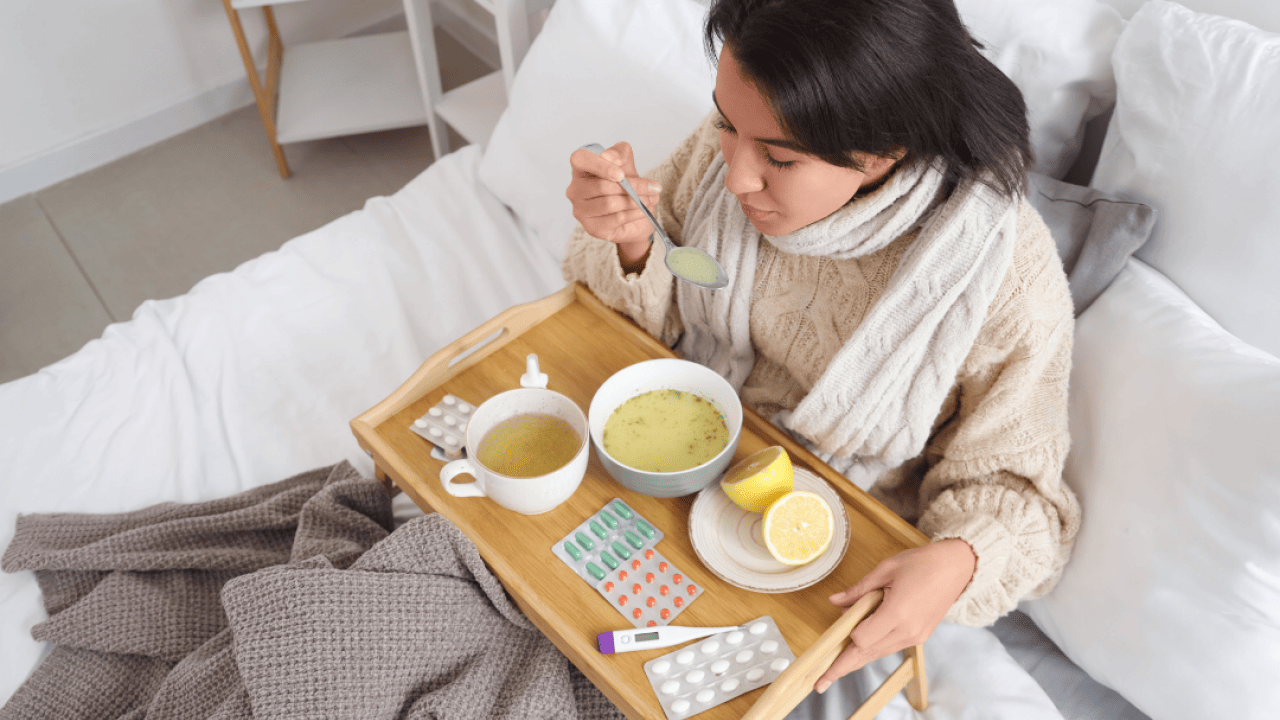 A woman sits in bed holding a spoon full of soup with a tray over her lap containing pills, nasal spray, a thermometer, lemons, a bowl of soup, and a mug of tea. 