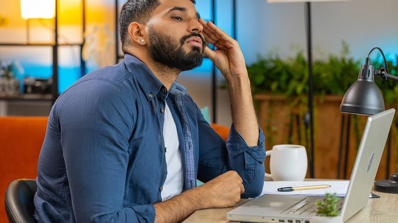 Man sitting at desk in modern colorful room looking at computer with an expression of distrust.
