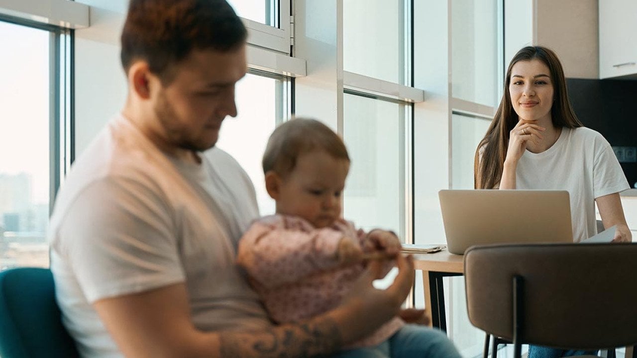 Woman smiling and sitting at table with laptop looking at man holding baby in the foreground.