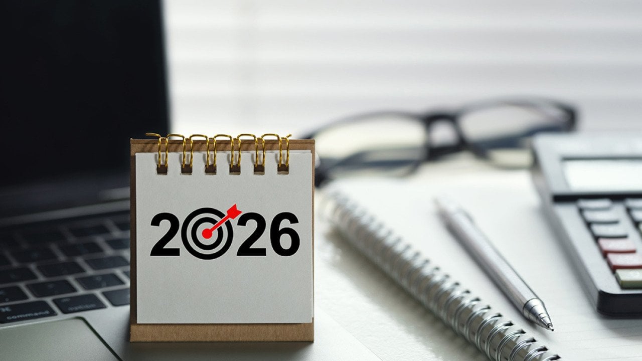 A desk calendar for 2026 with a target icon on top of a laptop among other office desk items.