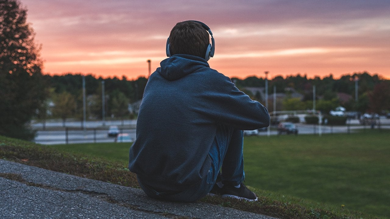Rear view of a male teenager sitting alone at the top of a hill and watching sunset while listening to headphones.