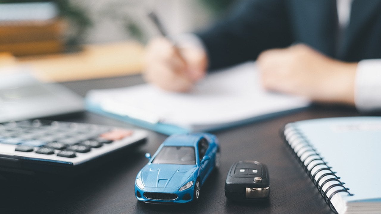 A small, blue car figure beside a car key in front of a car agent reviewing contracts.