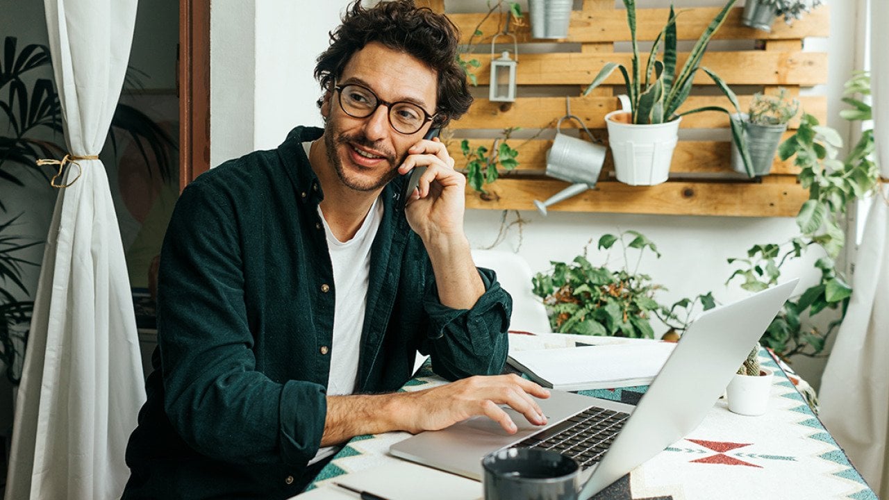 A young man on phone call and using laptop to work remotely from home.