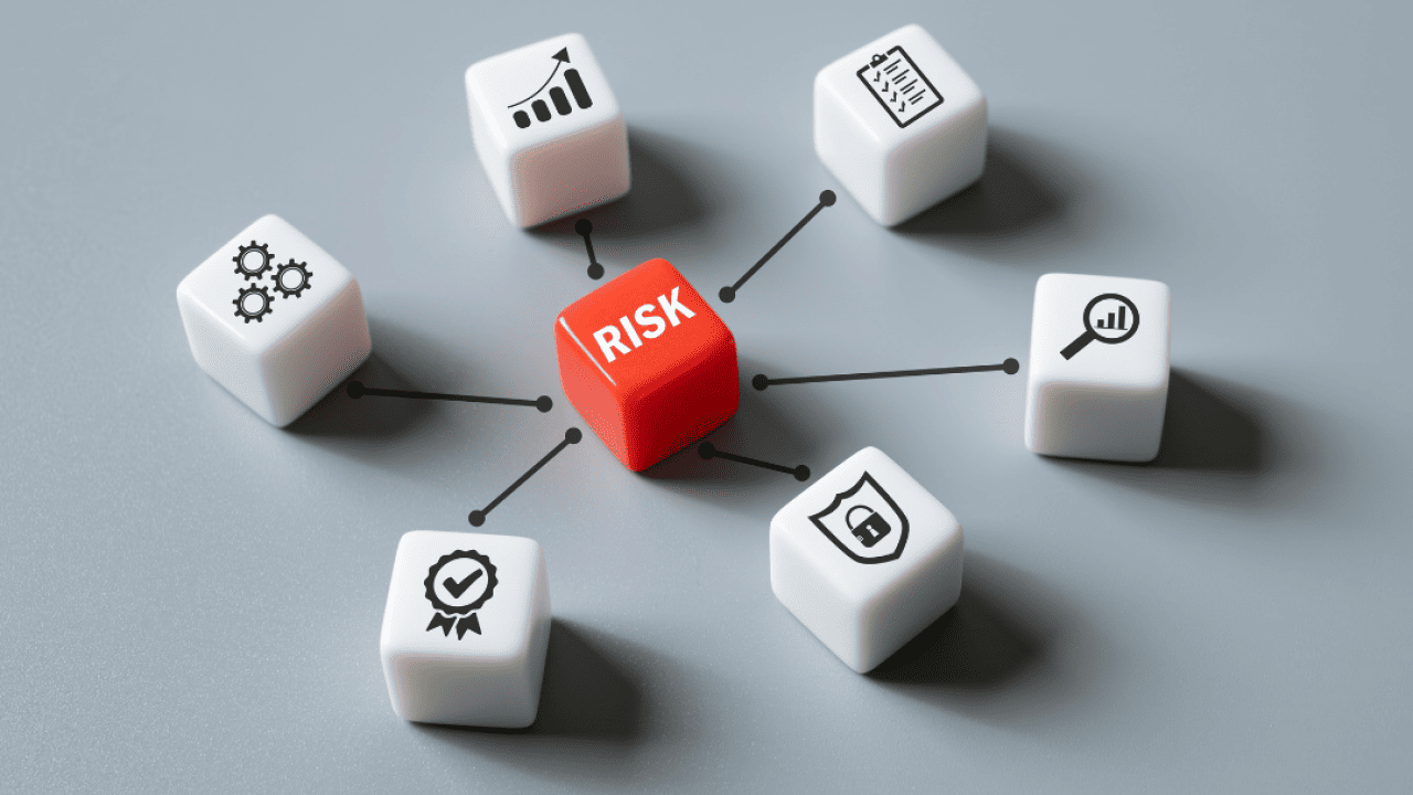 One red die with the word risk on it, positioned in in the middle of six white dice with symbols, including three cogs, an upward bar graph, a checklist, a magnifying glass over a rising bar graph, a security shield with a lock in the middle, and a ribbon with a check mark.
