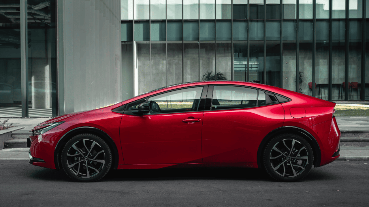 A red 2024 Toyota Prius Prime parked in front of a building on a city street.