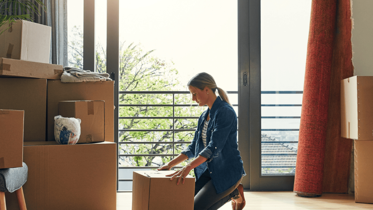 A women kneels in front of a cardboard box in a bright room with other boxes and household items like lamps and furniture. 
