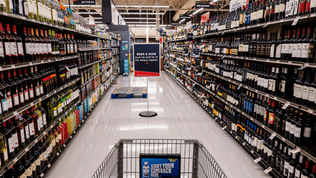 A shopping cart in a grocery store aisle filled with wine bottles.