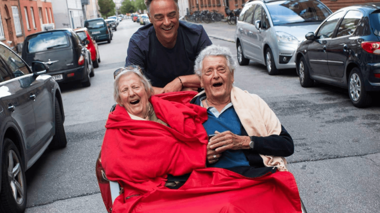 A volunteer cycles with a trishaw carrying two seated passengers who are laughing and smiling. 