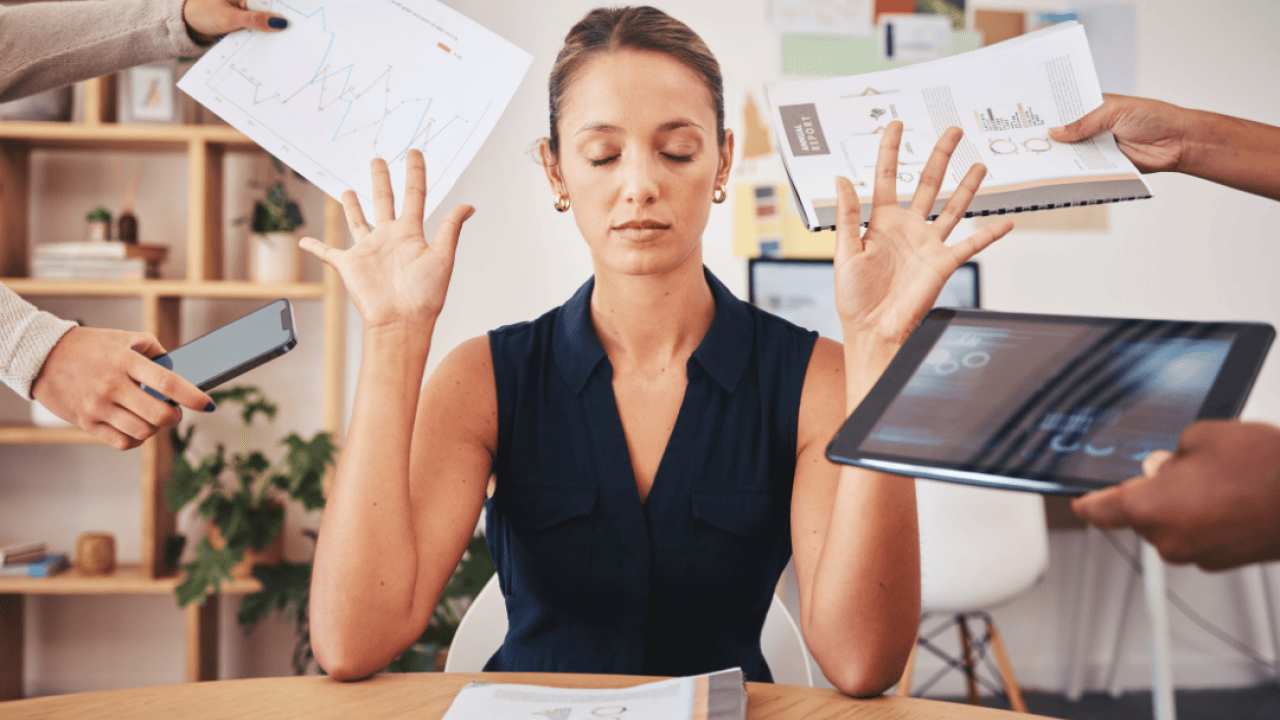 A women sitting in an office closes her eyes and puts her hands up as people hand her things including a phone, a computer, a paper with a graph on it, and a project notebook.