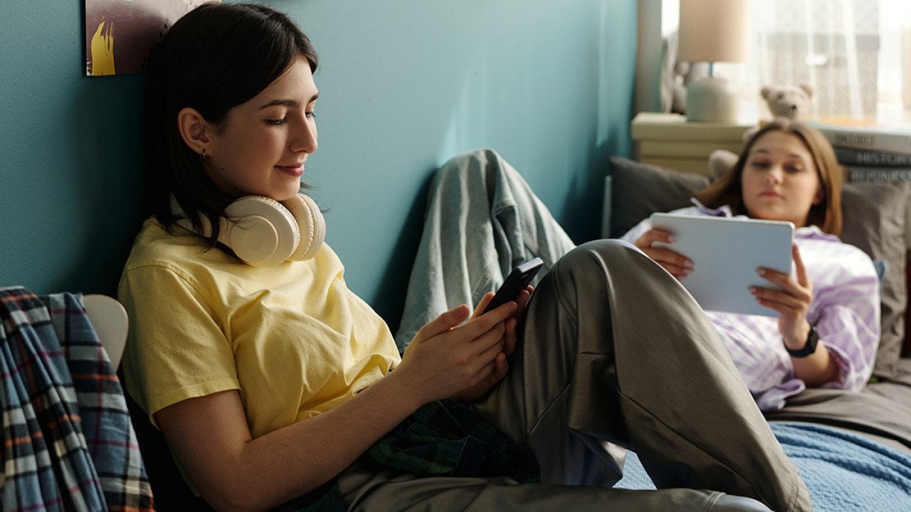 Two teenage girls relaxing on a bed and using smart devices to browse the internet.