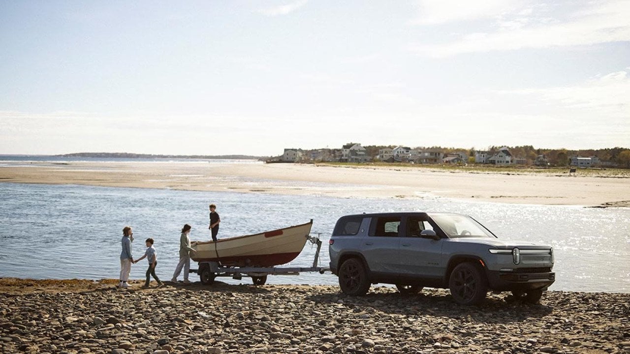 Rivian R1S towing a boat, parked near a body of water on a rocky dirt beach. A family of four plays near the boat.