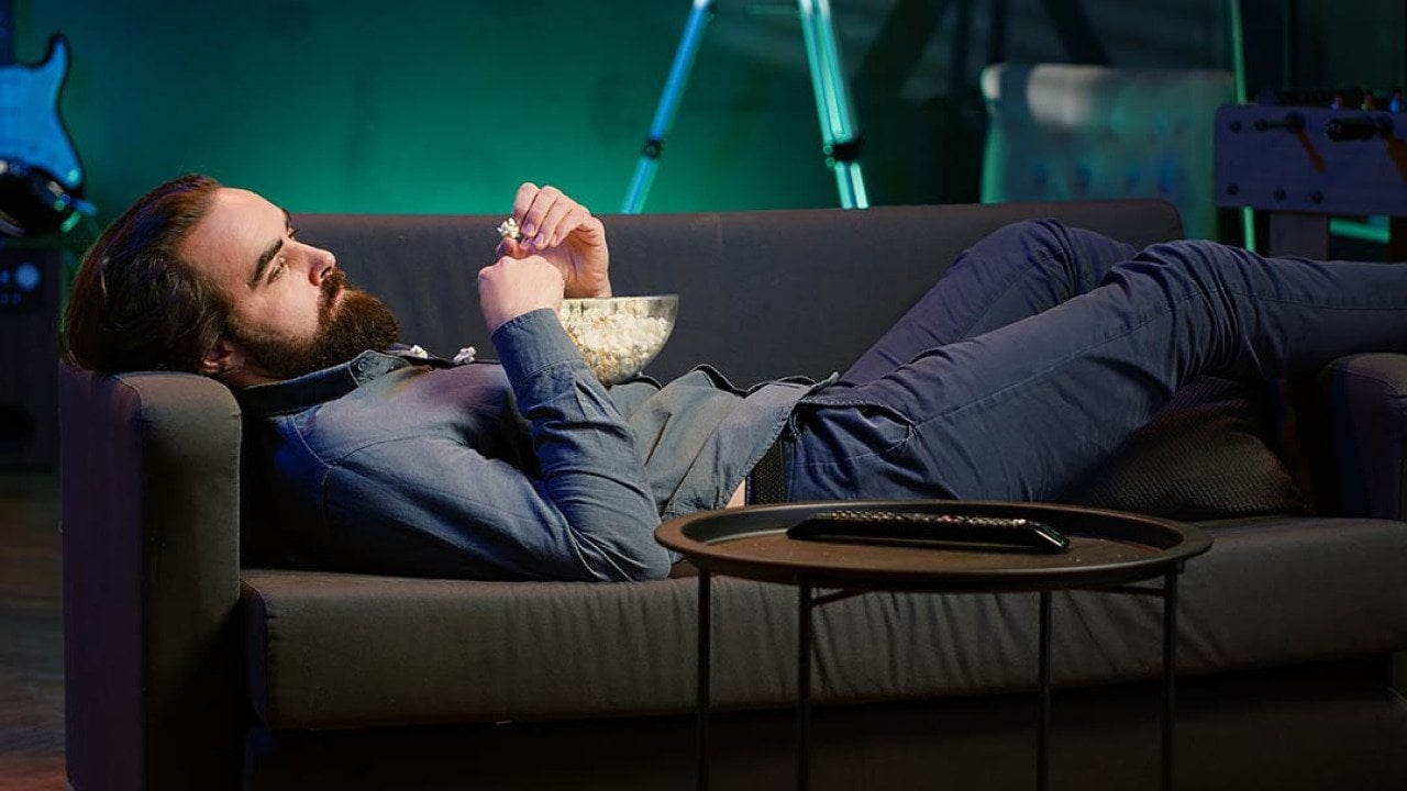 A bored man lying on couch and eating popcorn.