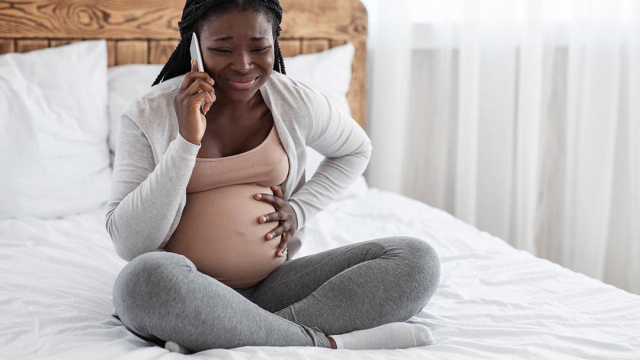 A pregnant young black woman calling on the phone while experiencing pain in her belly.