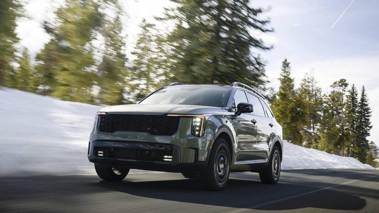 Low-angle view of earth-toned green 2026 Kita Sorento driving on mountain road with motion-blurred snow and pine trees in the background.