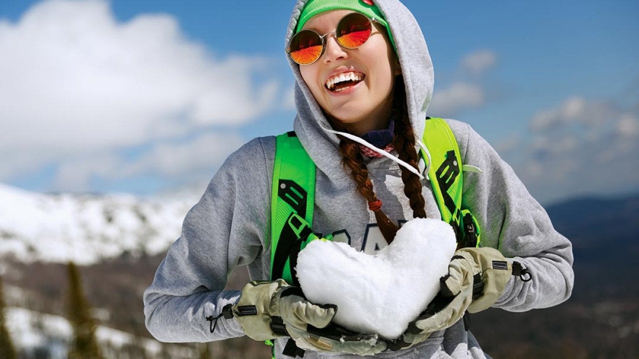 A young woman in snowboarding gear holding a heart shape made of snow.