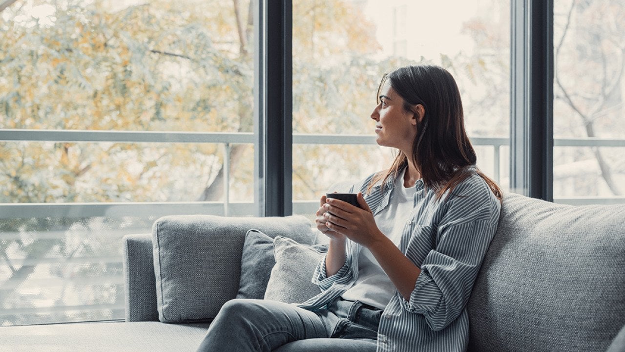 Young woman sitting on a sofa and having a cup of coffee while looking out the window at home.