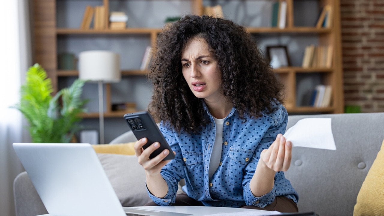 A young woman at home stressfully looking at her smartphone while calculating expenses.