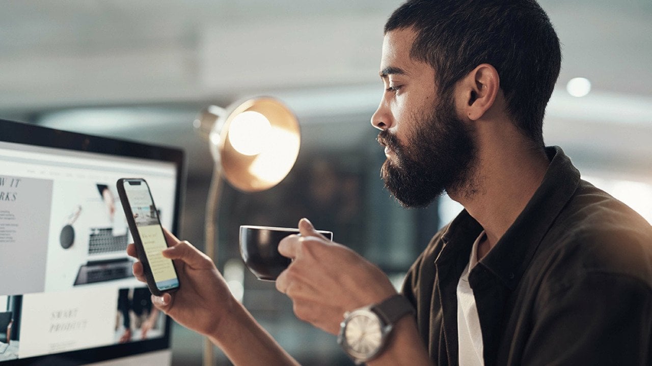 A man in a creative office checking social media on his phone while drinking a cup of coffee.