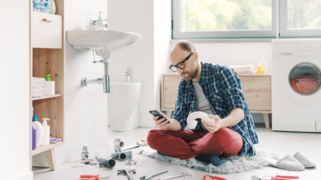 A man watching something on his smartphone while repairing a sink at home.