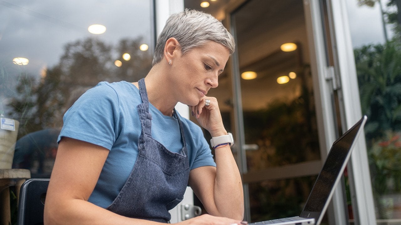 A female small business owner by the entrance of her store reviewing information on a laptop.