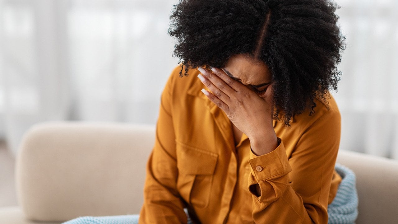 A young black woman sitting on a sofa holding her due to stress.