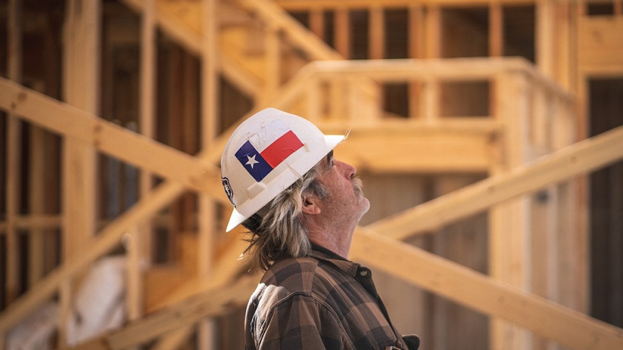 A construction worker wearing a hard hat with the Texas flag and looking up inside a wooden house construction site.