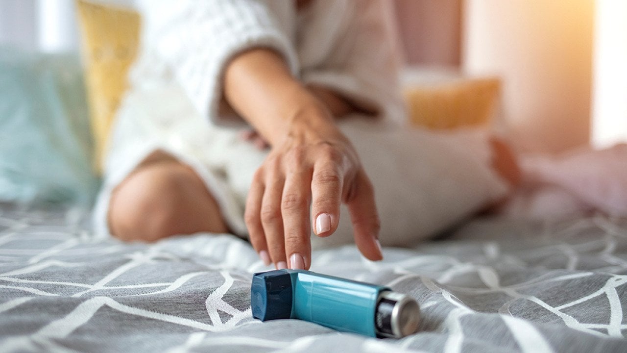 Woman sitting on a bed reaching for her inhaler.