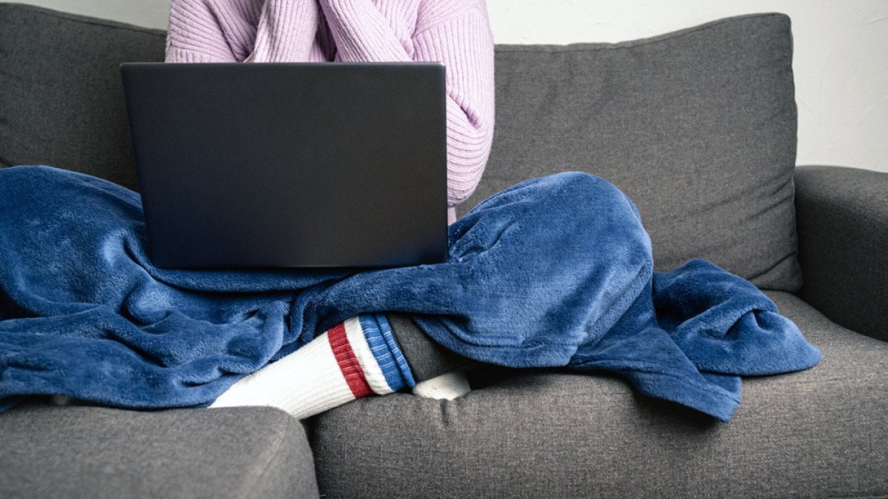 Person wearing a knitted sweater and socks with a blanket on while working and sitting on a sofa.