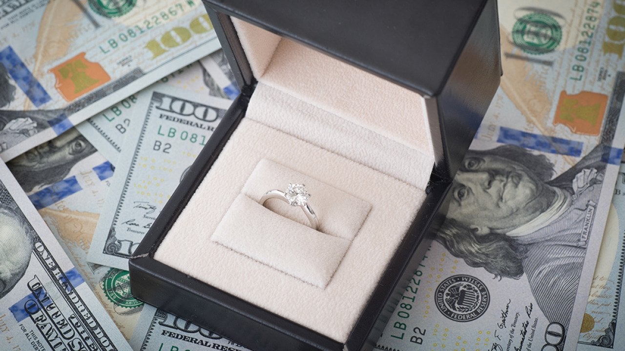 A diamond engagement ring placed above U.S. dollar bills.