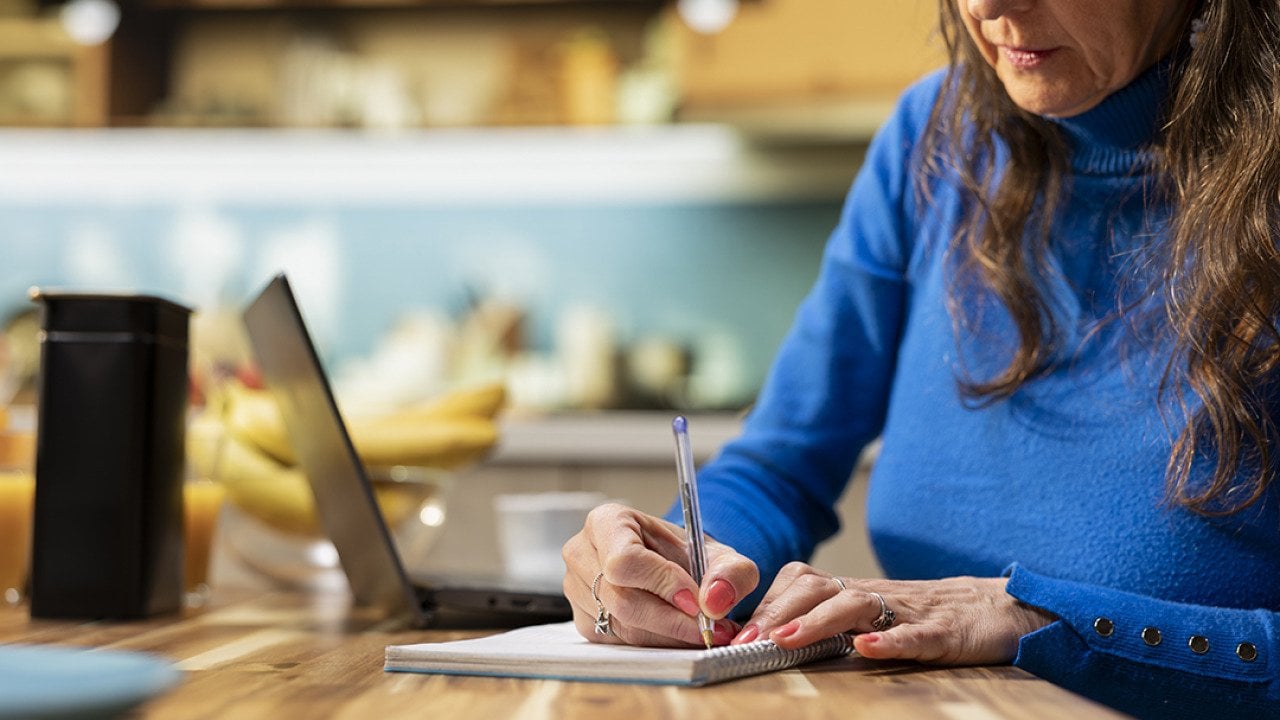 An elderly person writing a grocery list in a kitchen table.