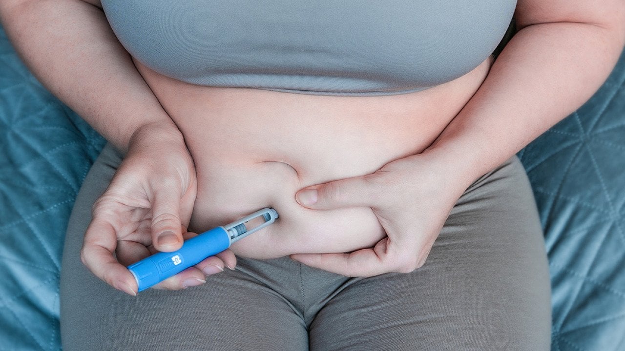 An overweight woman pinching belly fat to inject a GLP-1 pen on her stomach.