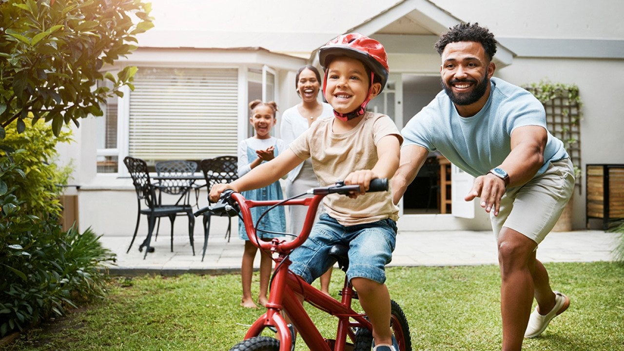 A young boy learning how to ride a bicycle being cheered on by his family.