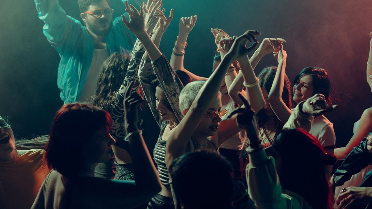 A crowd raising their hands as they dance under the lights in a club.