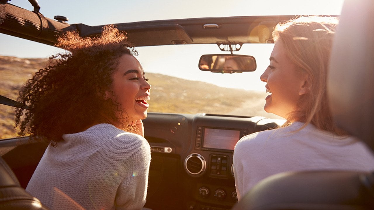 Two female friends happily riding on a highway in their convertible car.