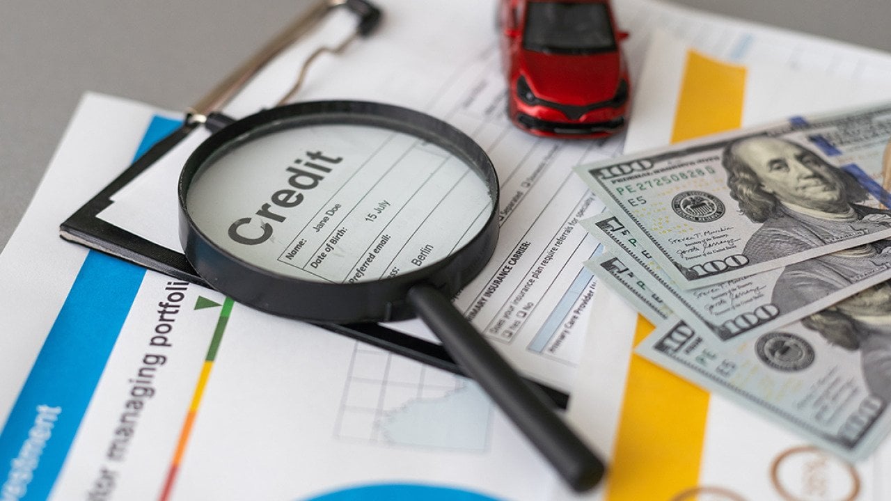 A credit loan application form with a magnifying glass, cash, and a small figure of a car on top of it.