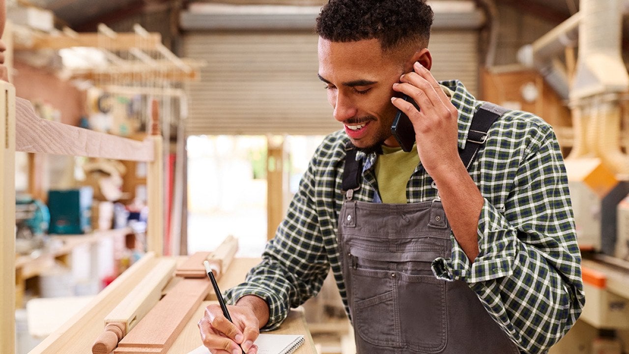 A carpenter taking a customer's order inquiry over a phone call in a furniture work shop.