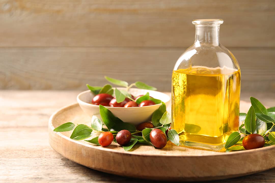 A glass bottle of jojoba oil with leaves and seeds of a shrub in a wooden plate.