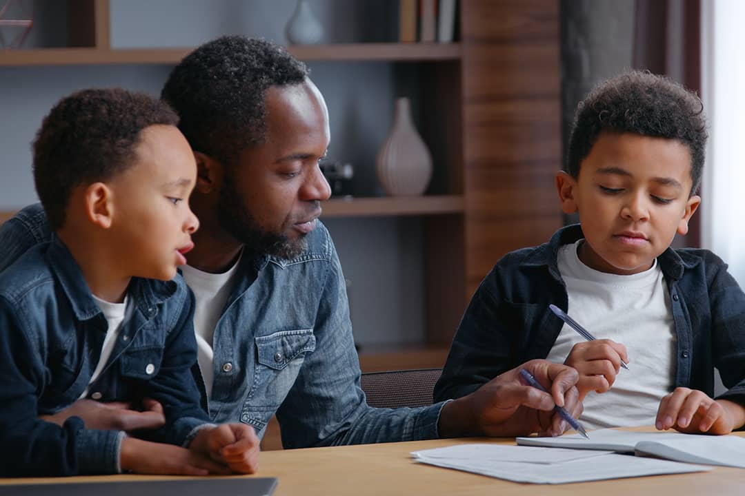 A parent studying with his young children, while helping his other son with a school writing assignment.