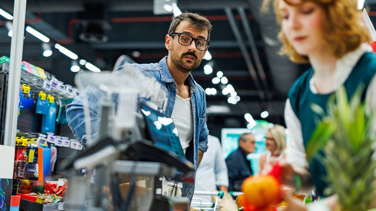A male customer wearing glasses and denim shirt watches the cashier scanning groceries at a checkout.