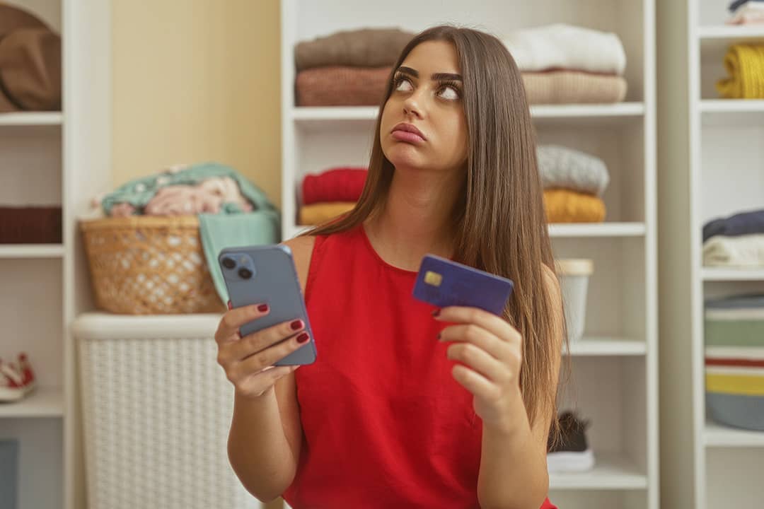 A young adult appearing to be frustrated while holding her smartphone and credit card in a dressing room.