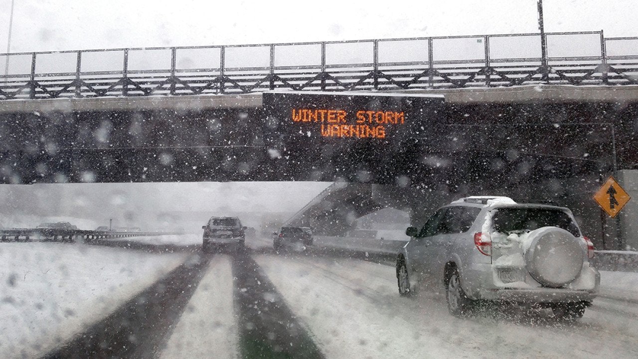 Windshield POV of a car passing under a bridge with an electronic 'Winter Storm Warning' sign.
