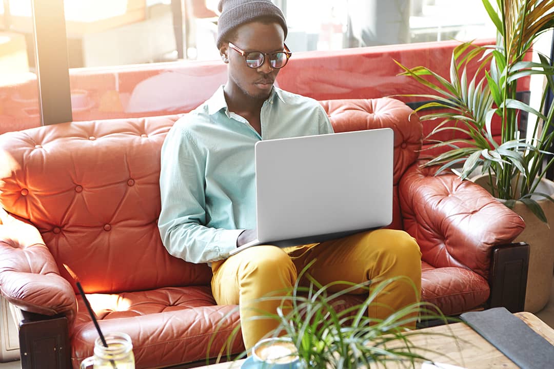A fashionable young black freelancer working remotely on a laptop while sitting on a red leather sofa.