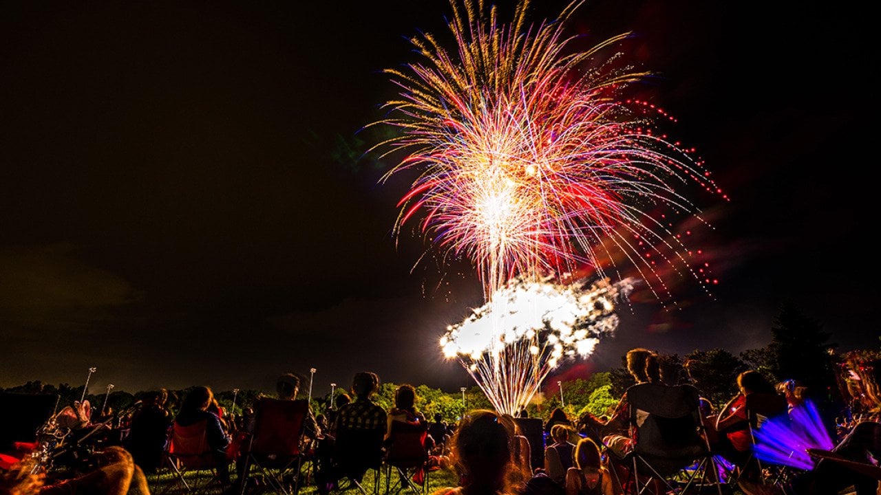 Crowds of people sit in lawn chairs and blankets to enjoy the colorful fourth of July independence day celebratory fireworks exploding in the black night sky.