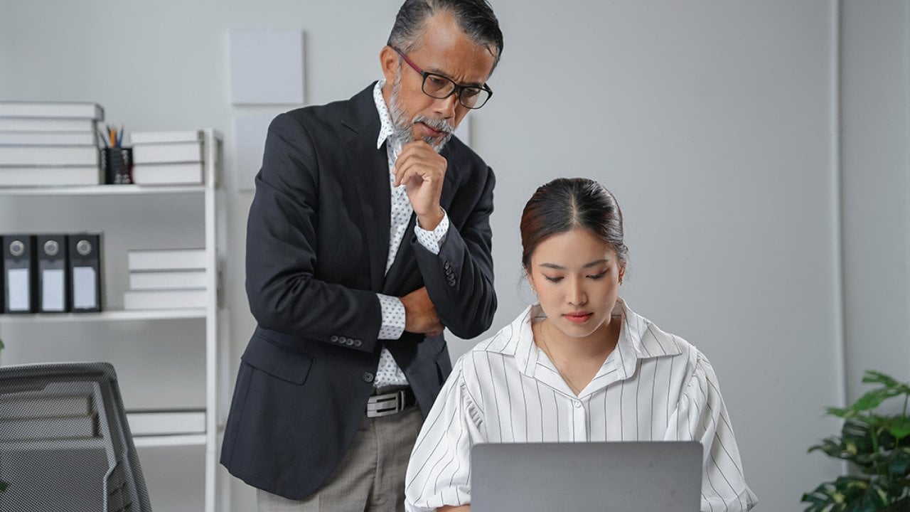 A manager standing behind an employee while checking her work report's progress.