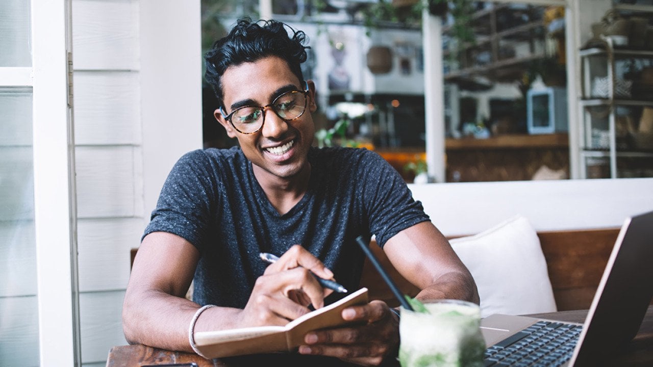 A happy young man taking notes while studying at a cafe.