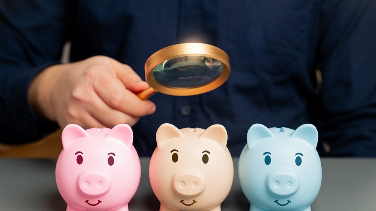 A person checking small piggy banks while using a magnifying glass, symbolizing analysis of savings.