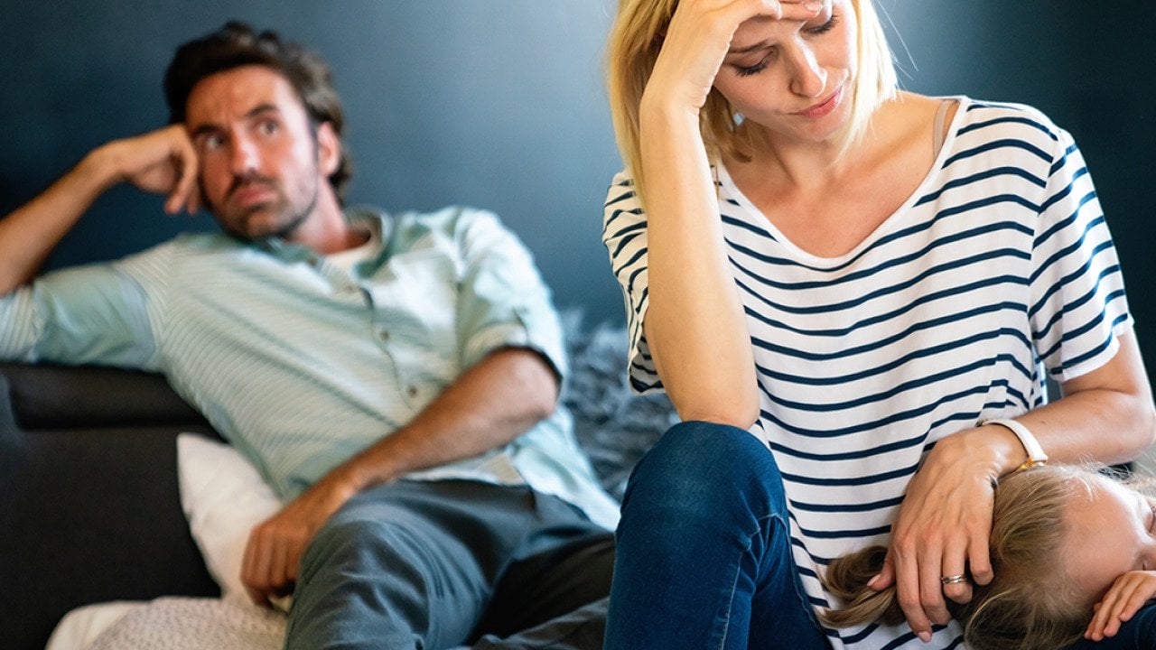 A stressed couple sitting in their living room while the mother comforts her young child.